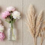 A symmetrically composed split-scene flat lay on a textured beige background, perfectly divided down the center. The left side displays fresh pink and white peonies, their soft petals full of moisture. The right side mirrors the composition with dried, feathery beige pampas grass plumes and delicate bunny tail grass, emphasizing textural contrast. The entire scene is bathed in soft, natural light, embodying a Pinterest home decor aesthetic.