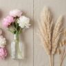 A symmetrically composed split-scene flat lay on a textured beige background, perfectly divided down the center. The left side displays fresh pink and white peonies, their soft petals full of moisture. The right side mirrors the composition with dried, feathery beige pampas grass plumes and delicate bunny tail grass, emphasizing textural contrast. The entire scene is bathed in soft, natural light, embodying a Pinterest home decor aesthetic.