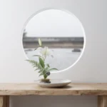 A complete minimalist ikebana arrangement with a white lily and green fern on a stone kenzan, centered on a light-wood table against a white wall with a circular window.