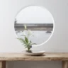 A complete minimalist ikebana arrangement with a white lily and green fern on a stone kenzan, centered on a light-wood table against a white wall with a circular window.